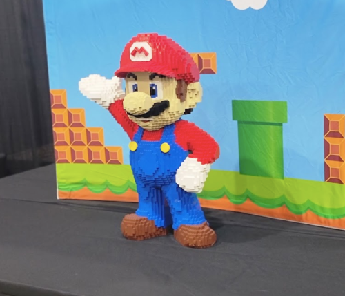 A LEGO sculpture of Mario waving, set against a colorful Super Mario background with pipes and blocks.