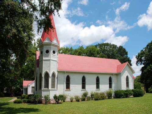 A white church with a red roof and steeple, surrounded by green grass and trees under a blue sky with clouds.