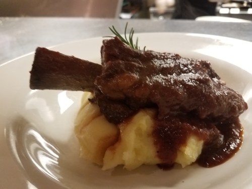 A plate of tender meat on creamy mashed potatoes, topped with rich gravy and garnished with a sprig of rosemary.