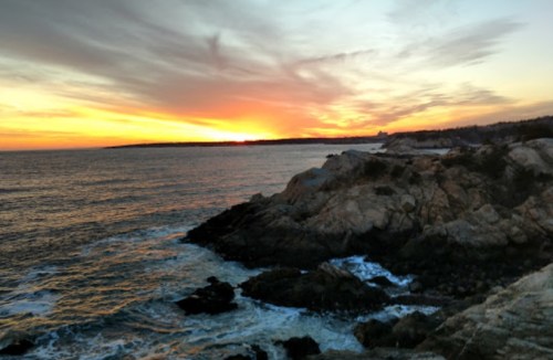 A serene coastal scene at sunset, with rocky shores and vibrant orange and purple skies reflecting on the water.