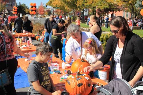 A lively outdoor event with families decorating pumpkins at tables, surrounded by autumn trees and festive decorations.