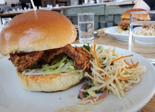 A close-up of a fried chicken sandwich with lettuce on a bun, served with a side of coleslaw and a glass of water.