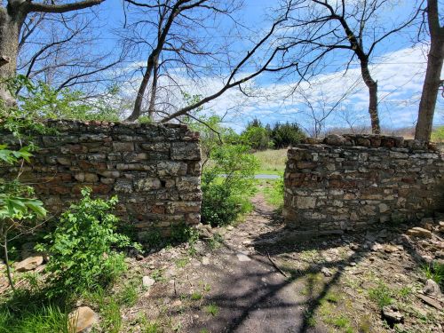 A stone wall with an opening, surrounded by trees and greenery, under a bright blue sky with scattered clouds.