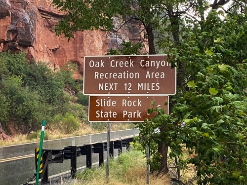 Sign indicating Oak Creek Canyon Recreation Area and Slide Rock State Park, next 12 miles, surrounded by greenery.