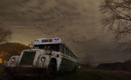 An old, weathered school bus sits in a field under a cloudy night sky, with trees in the background.