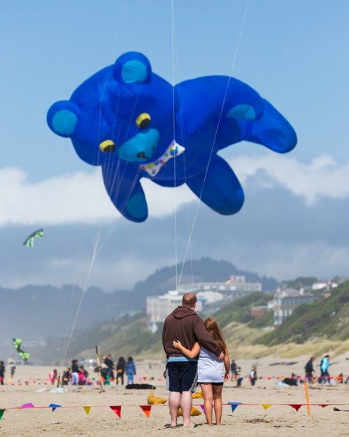 A couple watches a large blue bear kite flying above them at the beach, surrounded by colorful kites and people.