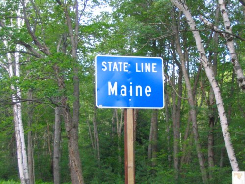 Blue state line sign marking the border of Maine, surrounded by green trees and foliage.