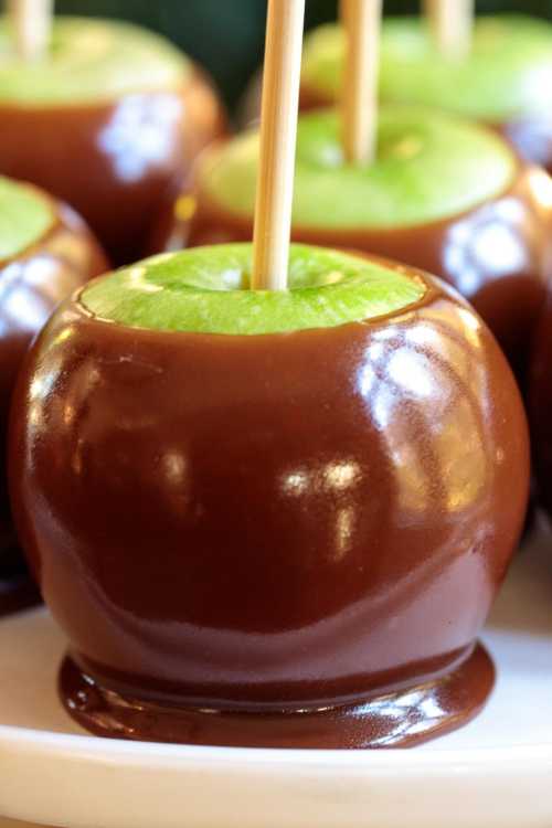 Close-up of shiny caramel-coated green apples on sticks, resting on a white plate.