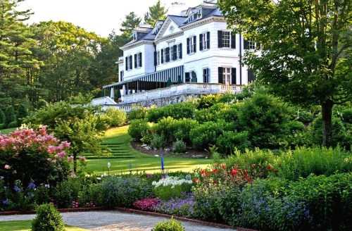 A grand white mansion surrounded by lush gardens and colorful flowers, set against a backdrop of trees.