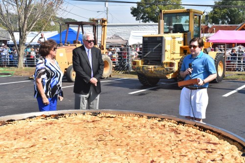 A large pie is displayed as a speaker addresses a crowd, with two people listening nearby and construction equipment in the background.