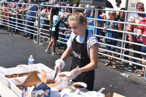 A young girl in an apron serves food to a long line of people at an outdoor event.