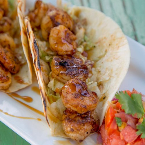 Three shrimp tacos on a plate, filled with shrimp, lettuce, and a side of fresh tomato salsa.