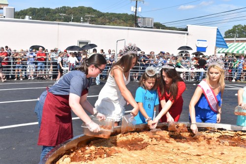 Participants prepare a large pie at a festival, with a crowd watching in the background.