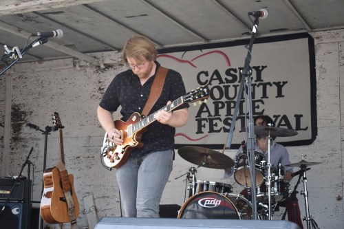 A musician plays guitar on stage at the Cass County Apple Festival, with a drummer in the background.