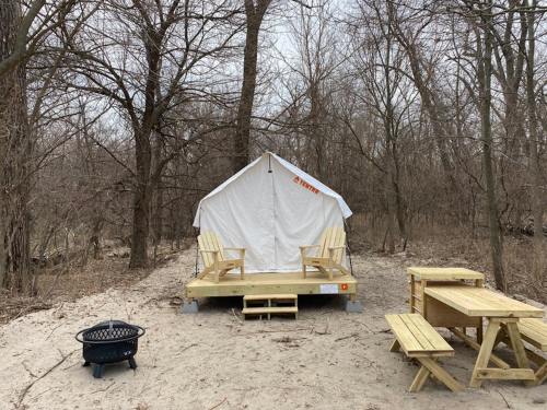 A white tent on a wooden platform in a wooded area, with picnic tables and a fire pit nearby.
