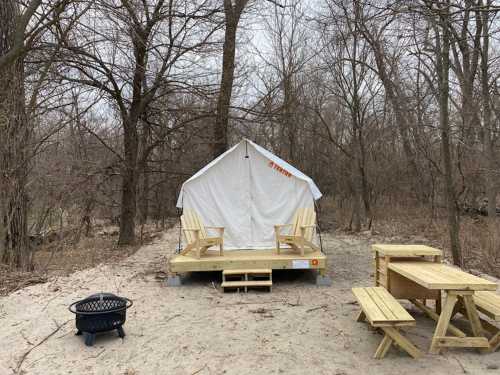 A white tent on a wooden platform in a wooded area, with picnic tables and a fire pit nearby.