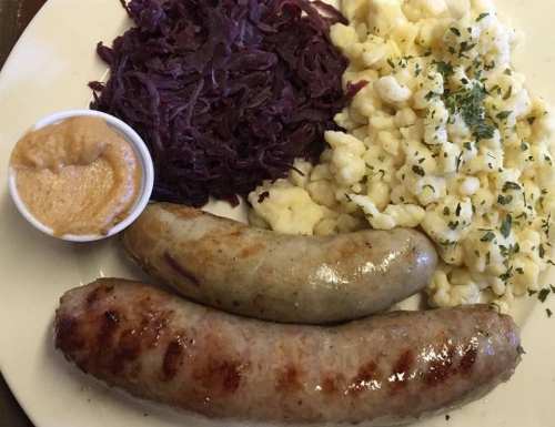 A plate with two sausages, red cabbage, creamy pasta, and a small cup of mustard.