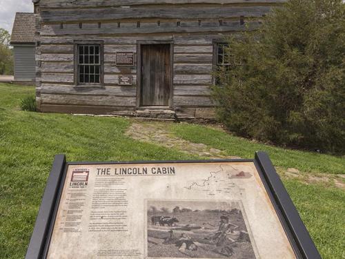 A historic log cabin with a sign in front, surrounded by grass and trees, representing the Lincoln Cabin.