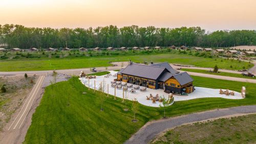 Aerial view of a modern lodge surrounded by green grass and trees, with outdoor seating and a winding road nearby.