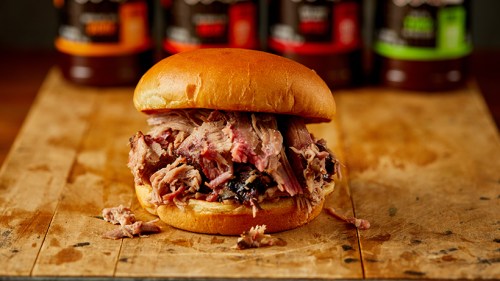 A pulled pork sandwich on a wooden board, with barbecue sauce bottles in the background.