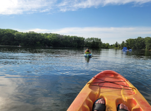 A view from a kayak on calm water, with another kayaker in the distance surrounded by greenery and blue sky.
