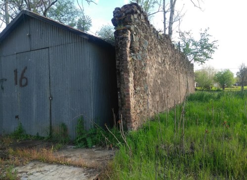 A weathered stone wall next to a metal shed, surrounded by overgrown grass and trees.