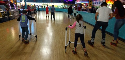 Two children use skating aids while roller skating in a colorful rink filled with people.