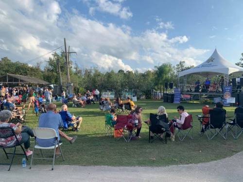 A crowd enjoys an outdoor event with a stage, seated on lawn chairs under a blue sky with scattered clouds.