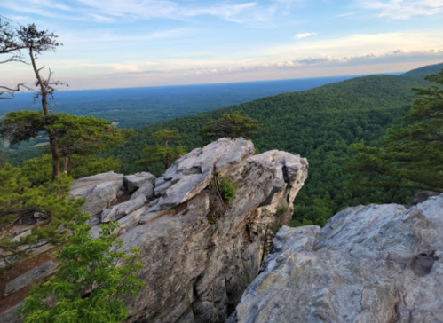 A rocky outcrop overlooks a lush green valley and distant mountains under a blue sky with scattered clouds.