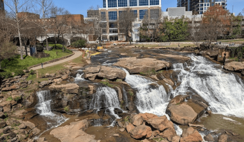 A scenic view of a waterfall cascading over rocks, with a park and buildings in the background on a sunny day.