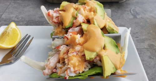 Two taco-shaped lettuce wraps filled with crab meat, avocado slices, and drizzled with a creamy sauce, served with a lemon wedge.