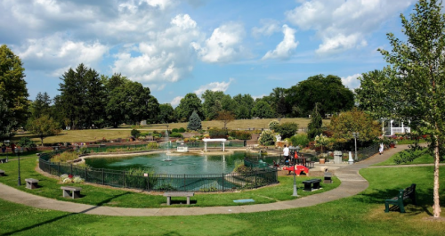 A serene park scene featuring a pond, walking paths, trees, and colorful flower beds under a partly cloudy sky.