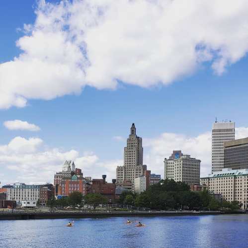 City skyline with modern buildings and a river in the foreground, under a partly cloudy sky.