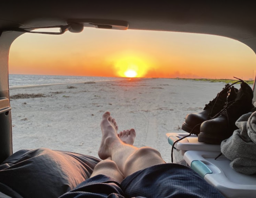 A person relaxes in a car, watching the sunset over a sandy beach, with boots and a towel nearby.