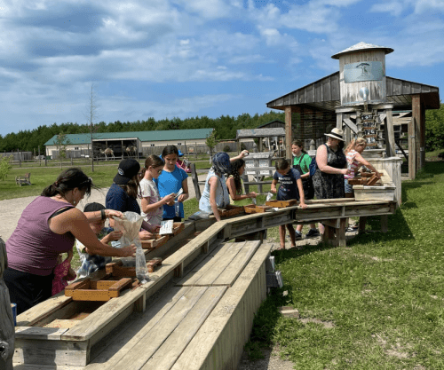 A group of children and adults are engaged in a hands-on activity at a wooden outdoor station, sifting through materials.