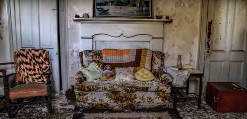 An old, worn sofa with colorful blankets and cushions, set in a peeling, vintage room with a fireplace and rustic decor.