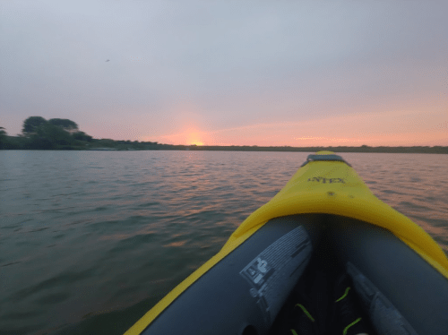 A view from a kayak on a calm lake at sunset, with vibrant colors in the sky and gentle ripples on the water.