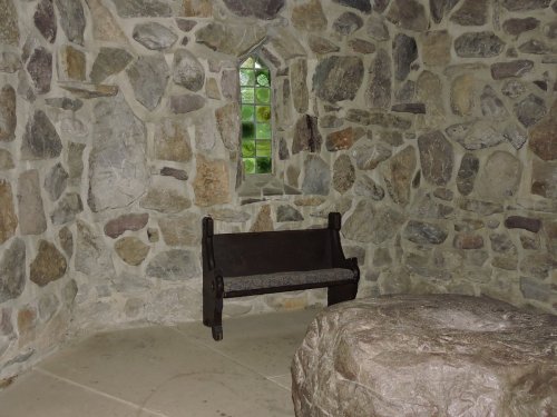 A stone room featuring a small wooden bench and a round stone table, with a window letting in natural light.
