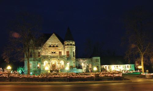 A beautifully lit stone mansion at night, adorned with festive lights and surrounded by trees.