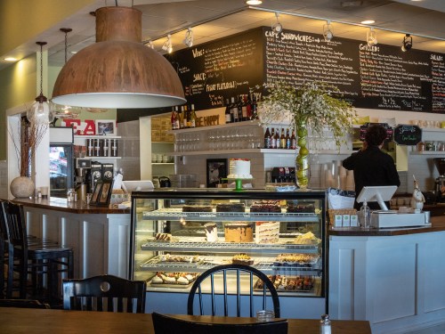 A cozy café interior featuring a display case of desserts, a counter with a barista, and a chalkboard menu.