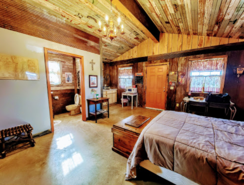 Cozy rustic bedroom with wooden walls, a bed, a small table, and a bathroom area, featuring warm lighting and decor.