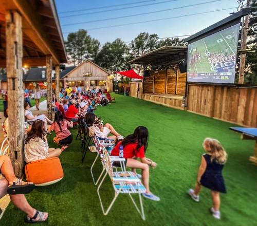A crowd watches a sports game on a large screen outdoors, seated on chairs in a grassy area with trees in the background.