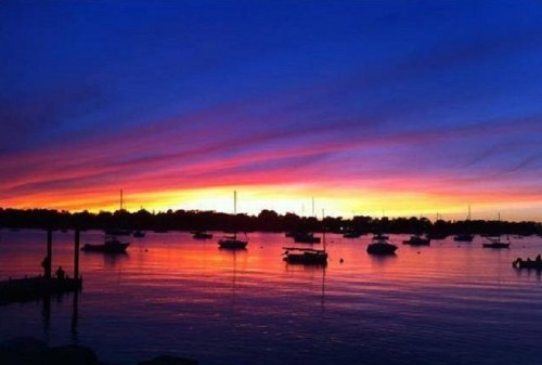 A vibrant sunset over a calm waterway, with silhouettes of boats and colorful clouds reflecting on the surface.