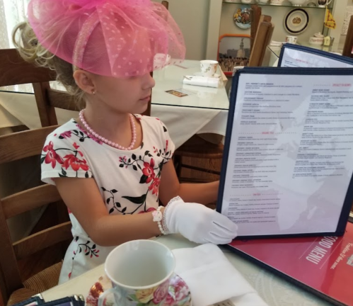 A young girl in a floral dress and pink hat examines a menu at a tea party, wearing white gloves and a pearl necklace.