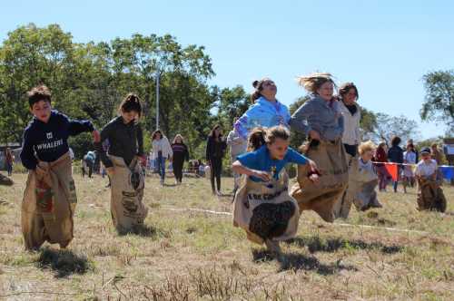 Children in burlap sacks race across a grassy field during a fun outdoor event on a sunny day.