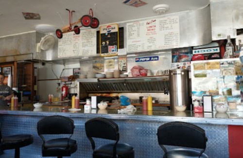 A retro diner interior with a metal counter, menus on the wall, and condiments on the tables.