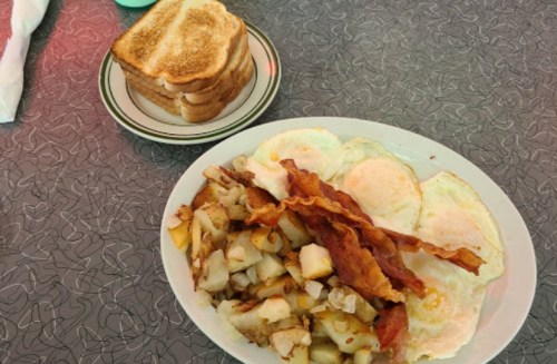 A plate of eggs, bacon, and hash browns next to a stack of toasted bread on a patterned table.