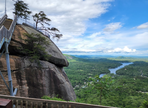A scenic view from a rocky overlook, featuring lush green hills and a winding river under a partly cloudy sky.