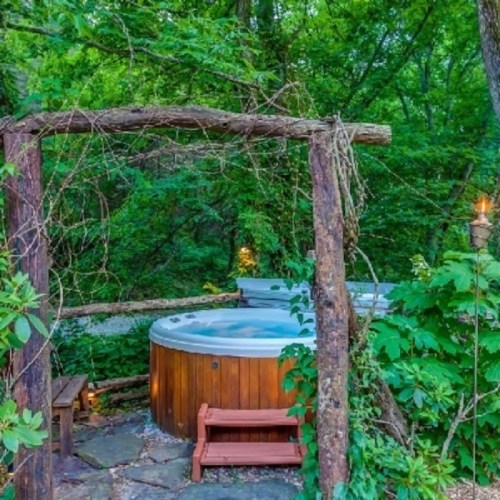 A wooden hot tub surrounded by lush greenery and rustic wooden structures in a serene outdoor setting.