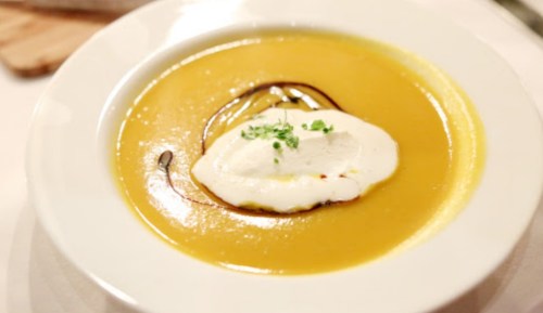 A bowl of creamy yellow soup topped with a dollop of sour cream and drizzled with olive oil, garnished with herbs.